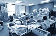 social worker drowing in files. There are files pilled up all over the desk and on top of filing cabinets. The pile of files practically reaches the ceiling.